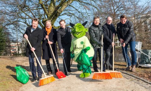 Sechs Männer stehen mit Besen und in der Mitte das Maskottchen "Grünhold".
